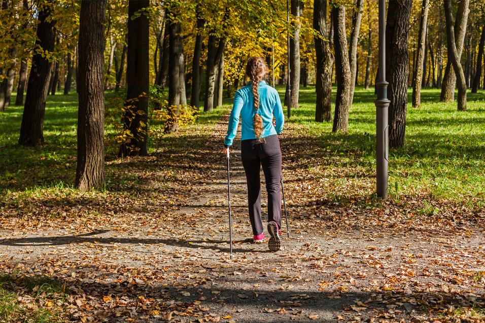 Frau mit langem Zopf geht mit Wanderstöcken einen herbstlichen Weg zwischen Bäumen, umgeben von buntem Laub.